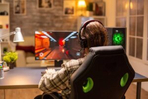 A gamer wearing headphones sits intently at a desk, focused on a colorful video game displayed on a large monitor.