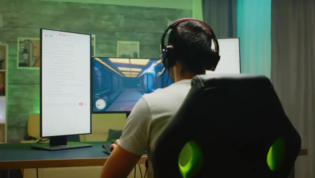 A gamer wearing headphones sits in a black chair, focused on dual monitors displaying a video game and a chat screen.
