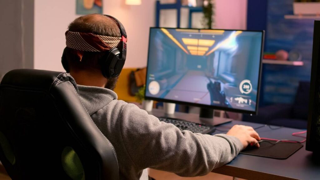 A man wearing headphones and a headband plays a video game, focused on the action displayed on his computer monitor.
