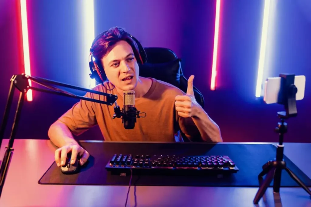 A gamer seated at a desk, wearing headphones, giving a thumbs up, with a microphone and keyboard in a colorful setup.