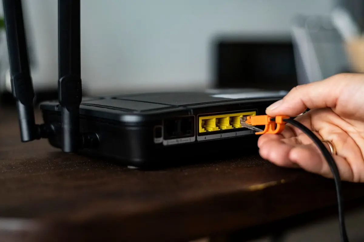 A hand connects an orange Ethernet cable to a black router with multiple yellow ports on a wooden table.