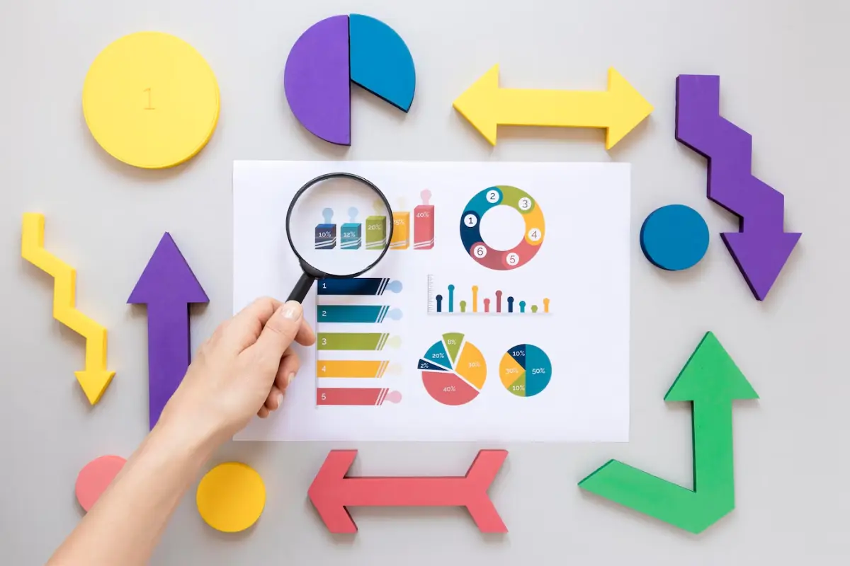 A hand holds a magnifying glass over colorful charts and graphs on paper, surrounded by various geometric shapes in vibrant colors.