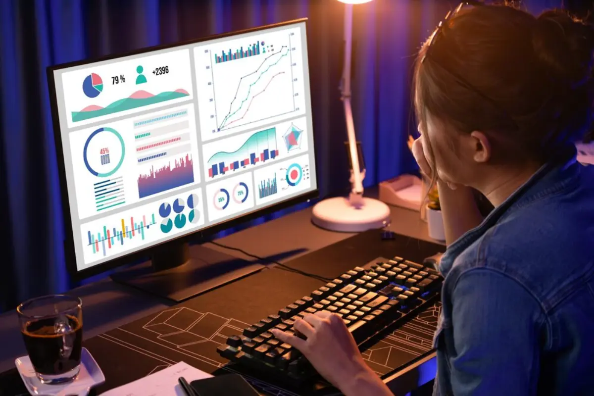 A person is analyzing colorful data visualizations on a computer screen, with a coffee cup and notepad nearby, in a dimly lit room.
