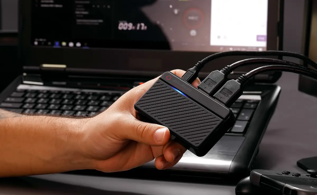 A person holds a black media capture device with multiple cables, in front of a laptop displaying a streaming interface.