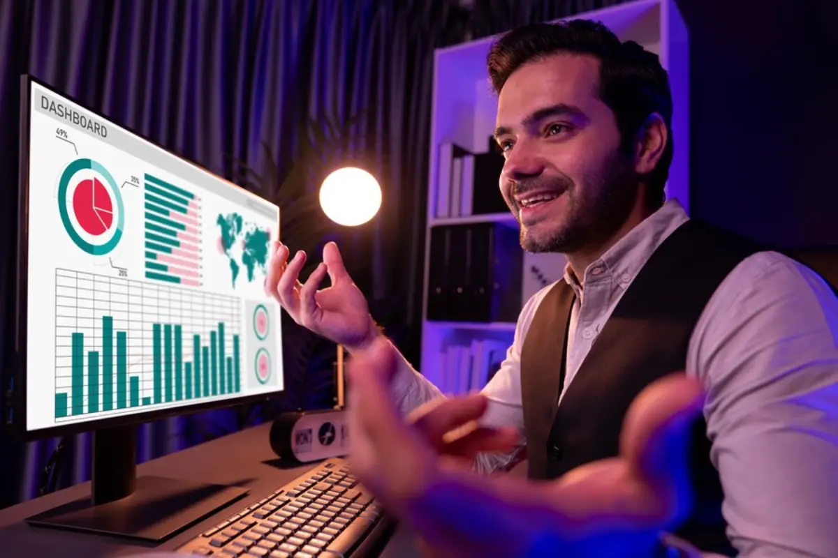 A person in a vest gestures towards a computer screen displaying colorful data visualizations and charts in a dimly lit room.