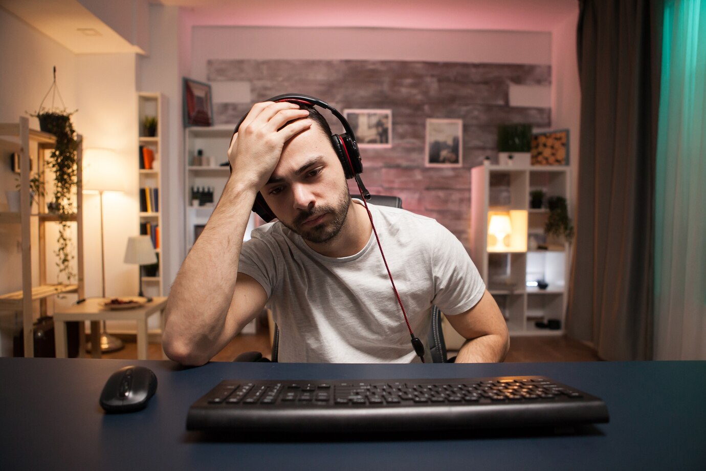 A person wearing headphones sits at a desk, resting their head in their hand, with a computer keyboard and mouse in front.