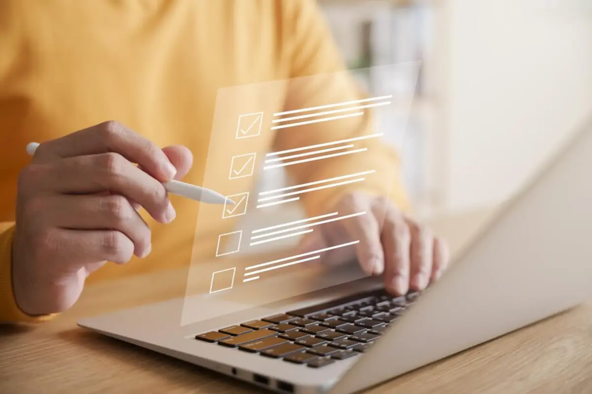 A person in a yellow sweater uses a stylus on a laptop, interacting with a digital checklist overlay.