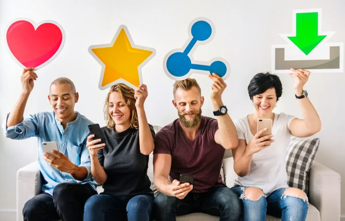 Four people sitting on a couch hold colorful social media reaction icons while looking at their phones.
