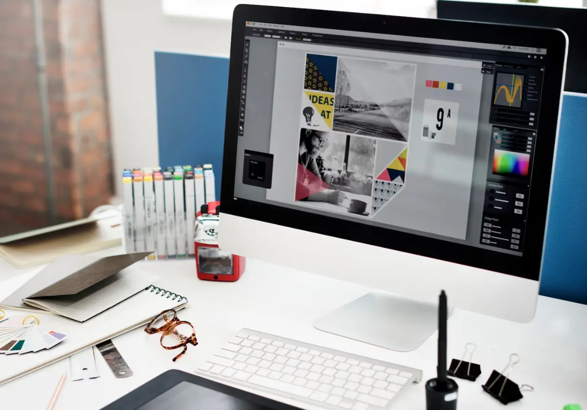 Image of a creative workspace featuring a computer displaying graphic design work, sketchbooks, pens, and color swatches on a desk.