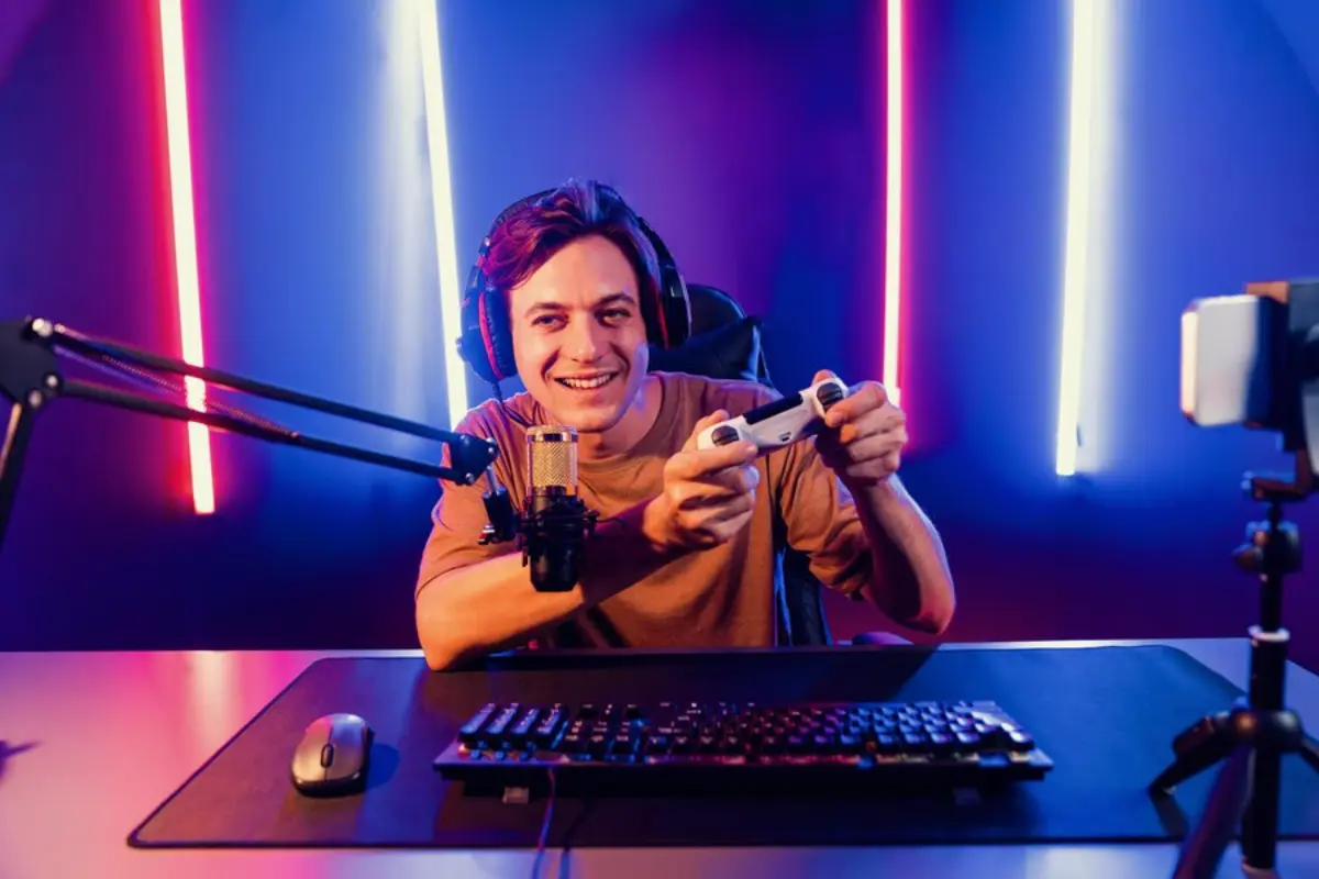 A gamer holds a white controller, wearing headphones and sitting at a desk with a microphone and keyboard, surrounded by colorful lighting.
