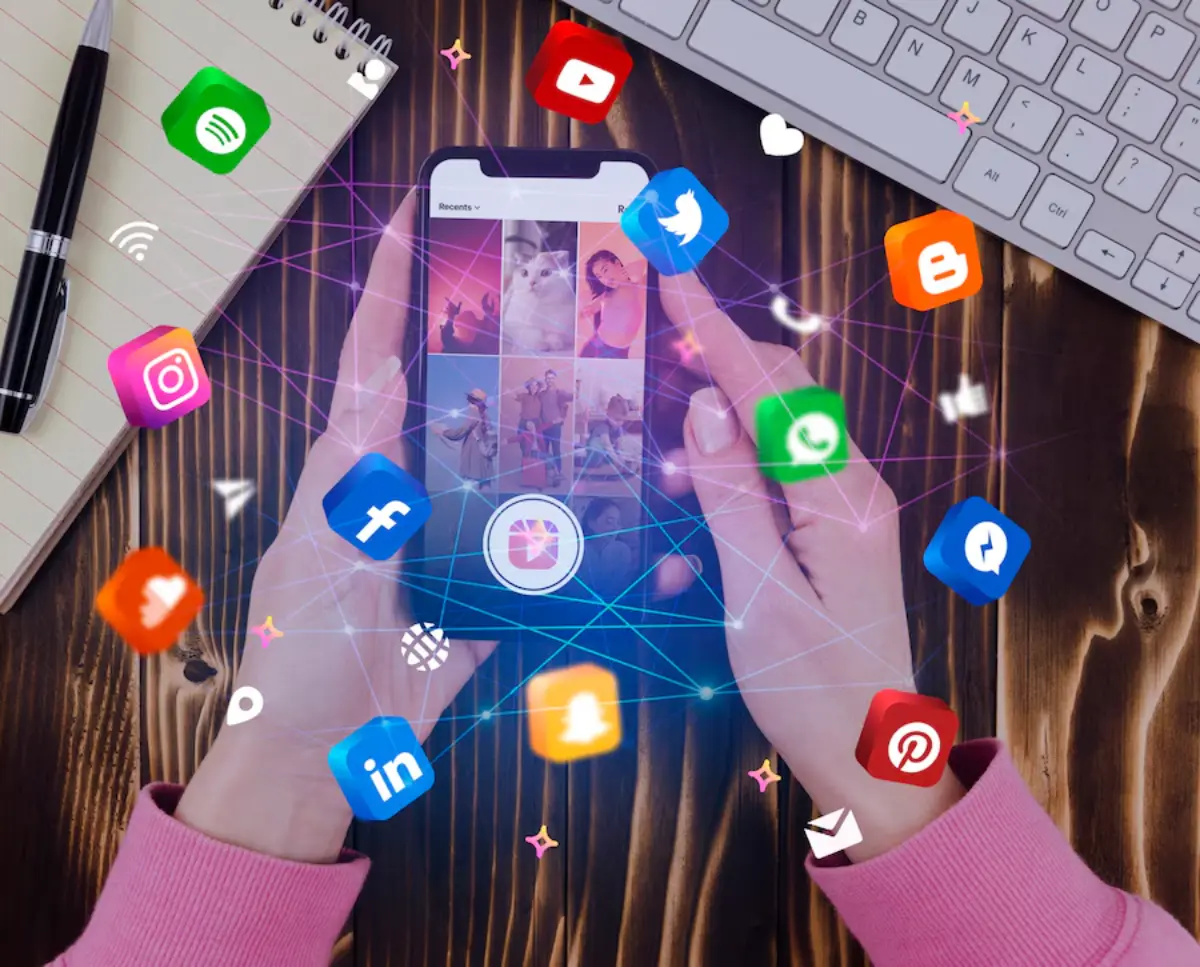 A person holds a smartphone displaying social media apps, surrounded by colorful app icons against a wooden background and notebook.