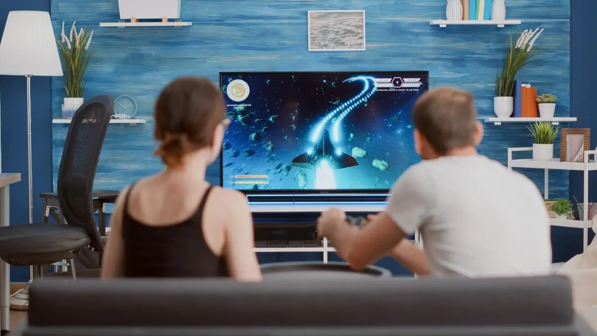 A couple sits on a couch, playing a space-themed video game on a large TV in a modern living room with blue accents.
