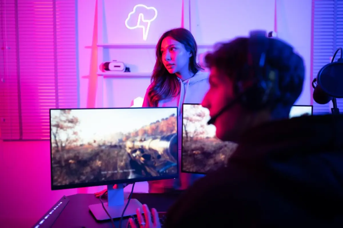 A gamer sits at a dual monitor setup, illuminated by colorful lights, focusing intently on a video game landscape.
