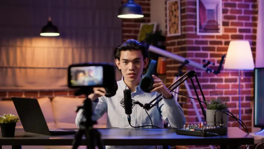 A person sits at a desk in a cozy, brick-walled room, speaking into a microphone, with a laptop and recording equipment in view.