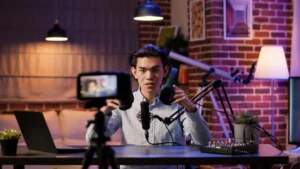 A person sits at a desk in a cozy, brick-walled room, speaking into a microphone, with a laptop and recording equipment in view.