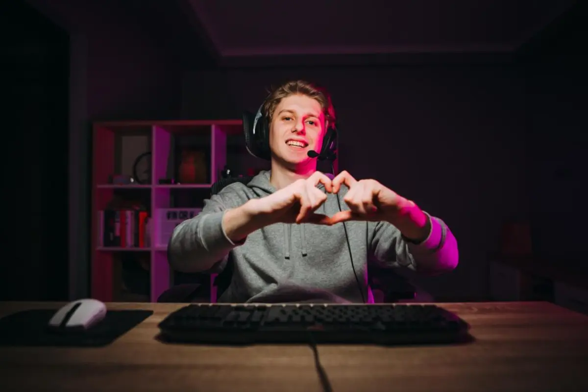 A gamer in a headset makes a heart shape with hands, sitting at a wooden desk with a keyboard and mouse, illuminated by pink light.