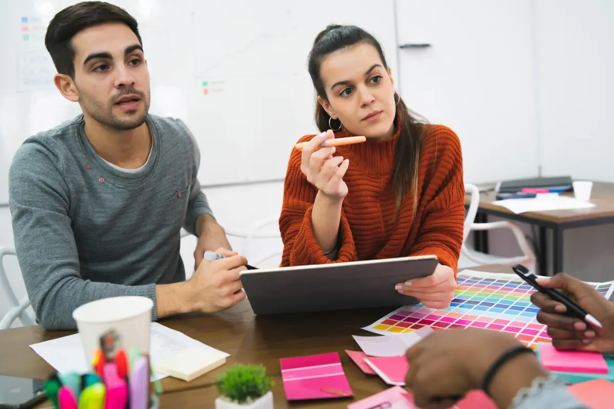 A group of people collaborates around a table, discussing ideas while surrounded by colorful design samples and stationery.