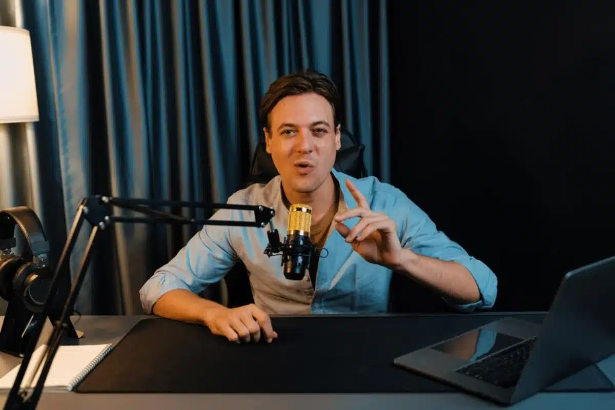 A person in a light blue shirt gestures while sitting at a desk with a microphone, laptop, and headphones against a dark backdrop.