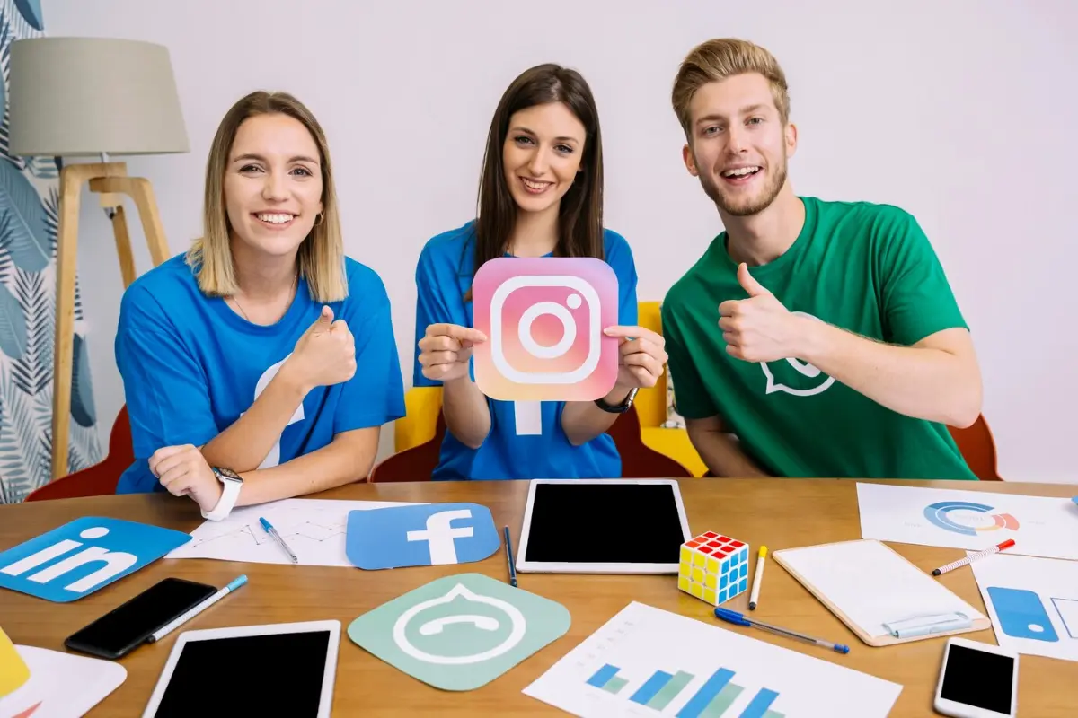 Three individuals hold social media logos, including a large Instagram icon, surrounded by tablets and marketing materials on a table.