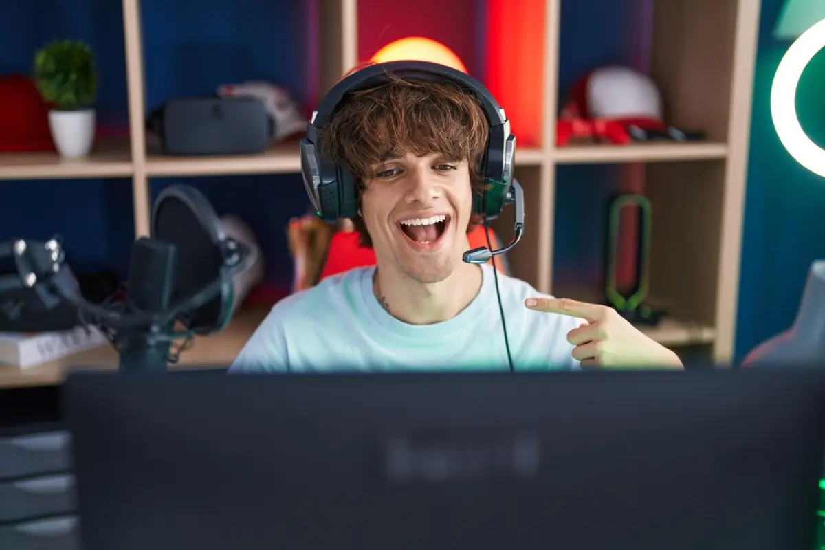 A person wearing a headset points at a computer monitor in a colorful gaming setup, surrounded by shelves and equipment.