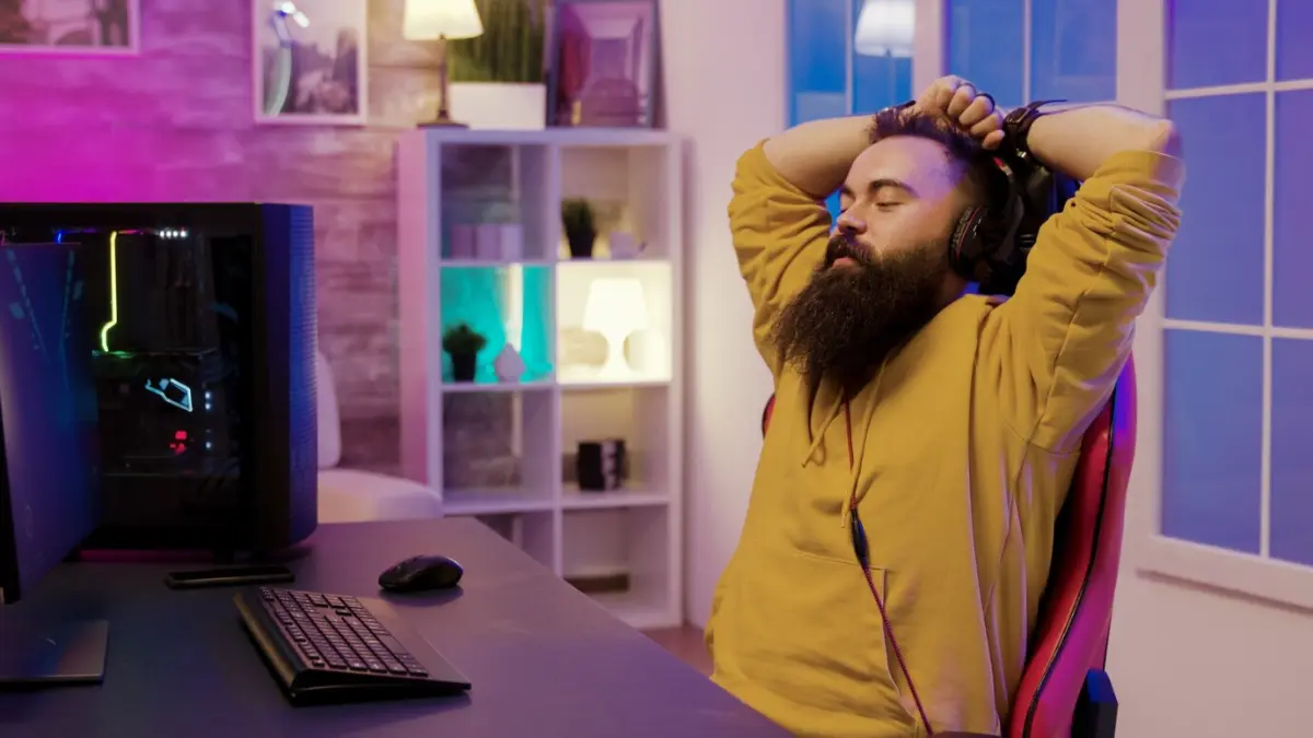 A gamer in a yellow hoodie leans back in a red gaming chair, with a colorful gaming setup and soft lighting in a cozy room.