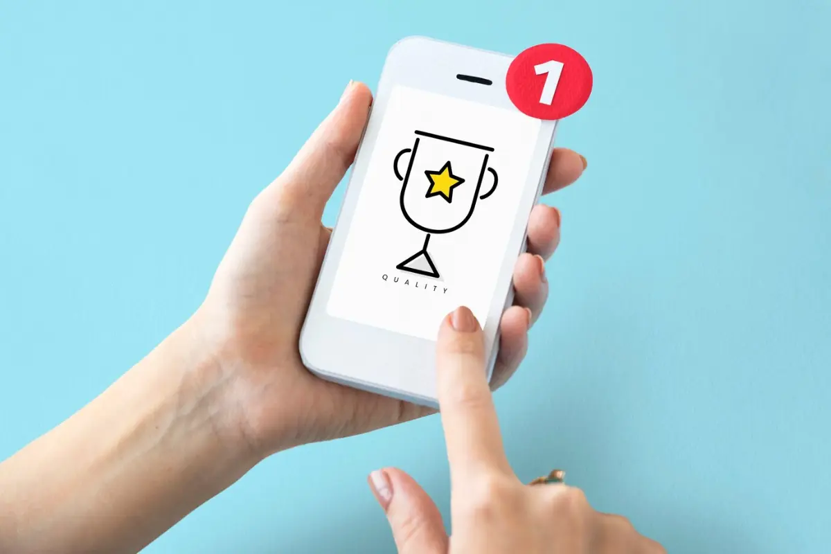 A hand holds a smartphone displaying a trophy icon with a yellow star and the word QUALITY, accompanied by a notification badge.