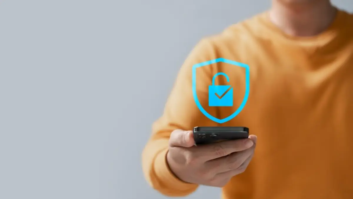 A person in a yellow sweater holds a smartphone, with a blue shield and lock icon symbolizing digital security hovering above it.