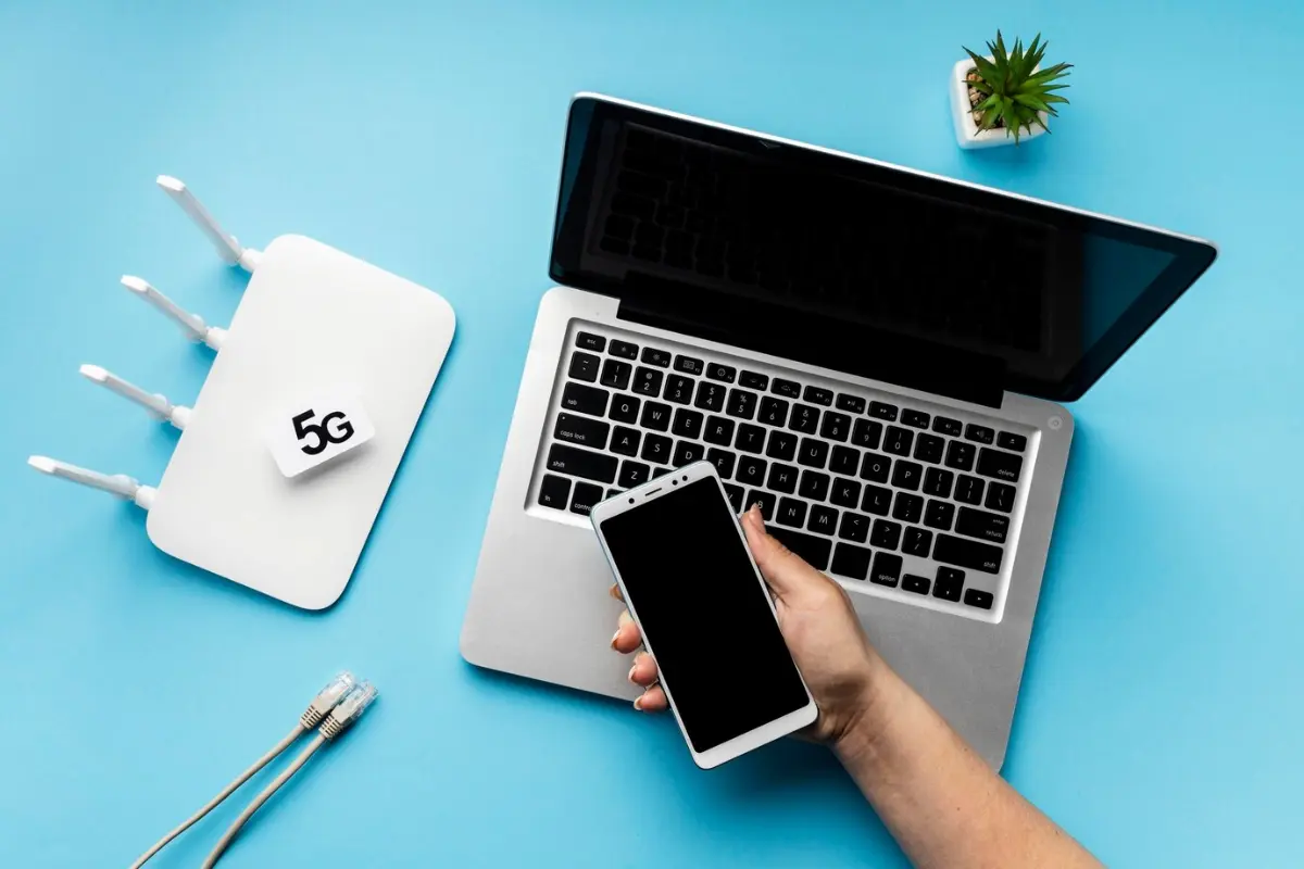 A hand holds a smartphone next to a laptop and a 5G router on a blue background, with Ethernet cables nearby.