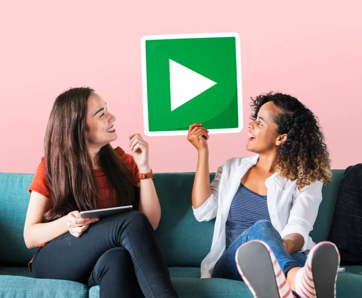 Two women sit on a teal couch, one holding a large green play button sign, against a soft pink background. One uses a tablet.