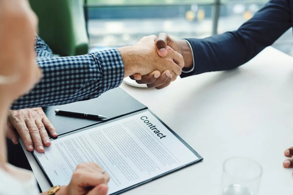 Two individuals shake hands over a contract on a table, symbolizing agreement and partnership in a professional setting.