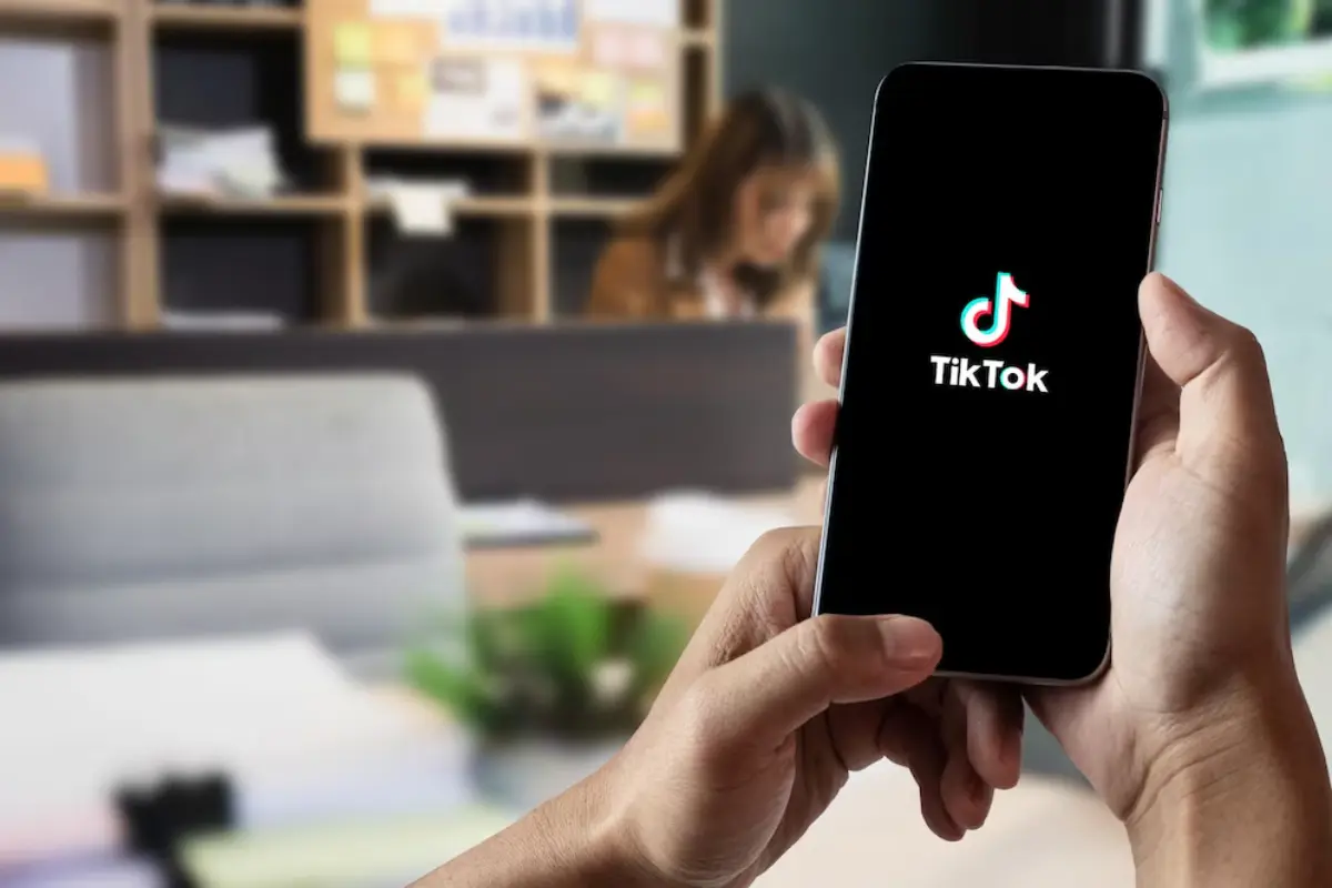 A person holds a smartphone displaying the TikTok logo, set in a modern office environment with shelves and plants in the background.