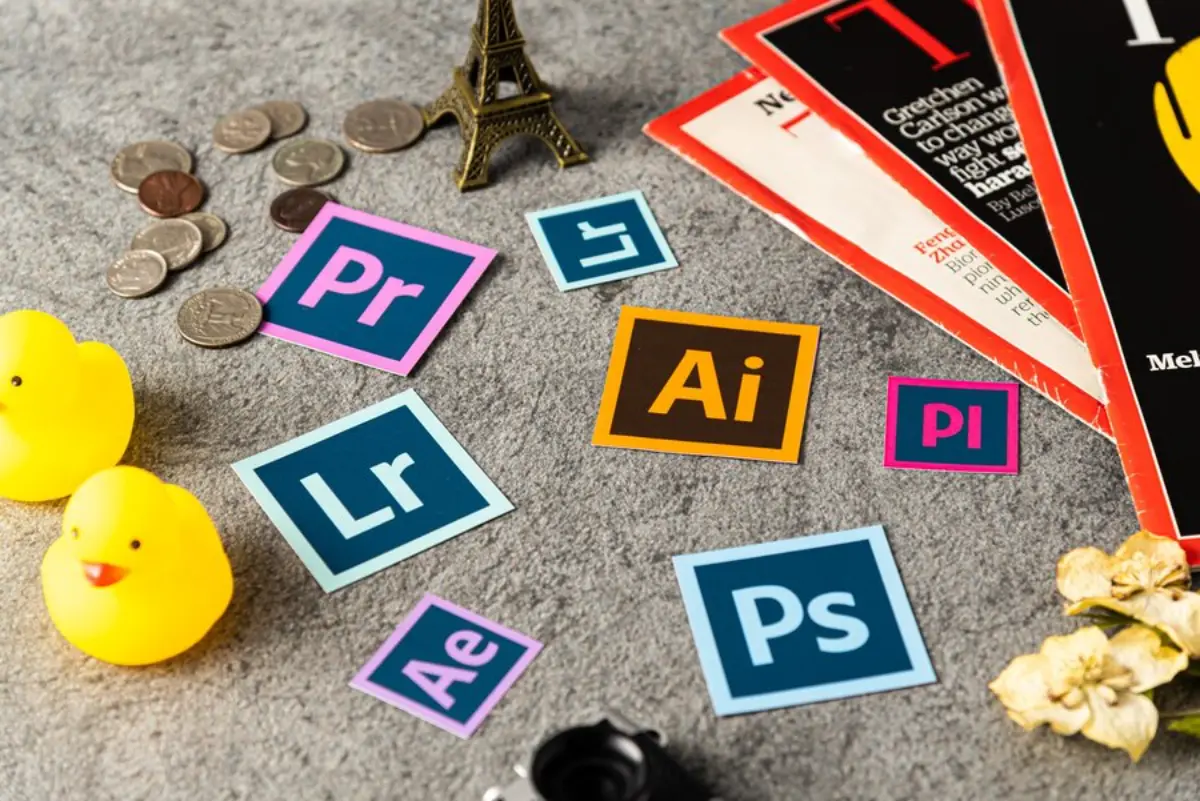 Colorful Adobe icons on a textured surface with coins, a mini Eiffel Tower, rubber ducks, and magazines, showcasing creative tools.