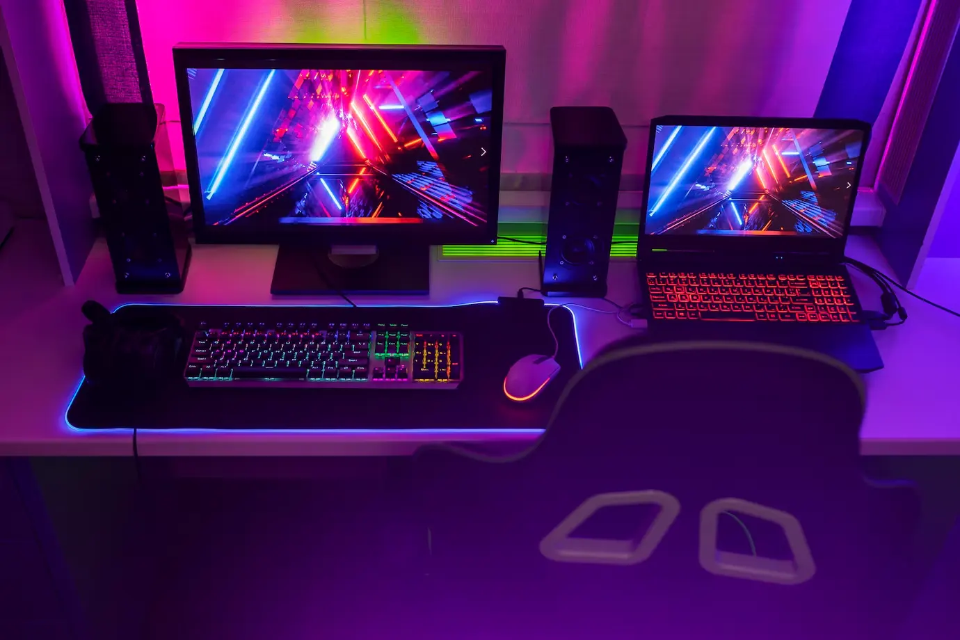 A vibrant gaming setup featuring two screens, a colorful keyboard, mouse, and neon lighting, on a sleek minimalist desk.