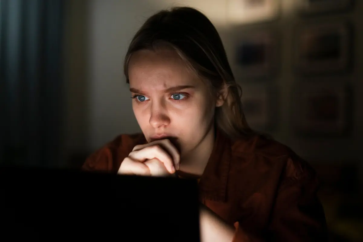 A person sitting in a dimly lit room, focused on a laptop, with soft shadows highlighting their features and surroundings.