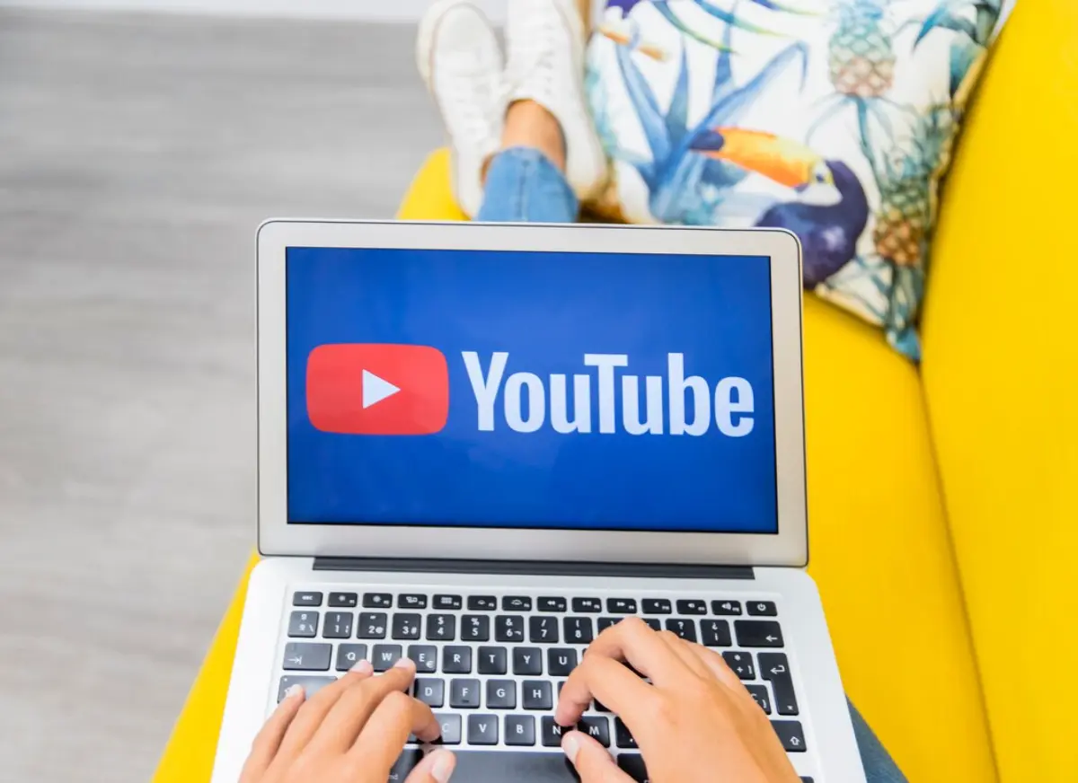 A person using a laptop with the YouTube logo on the screen, sitting on a yellow sofa with colorful cushions.