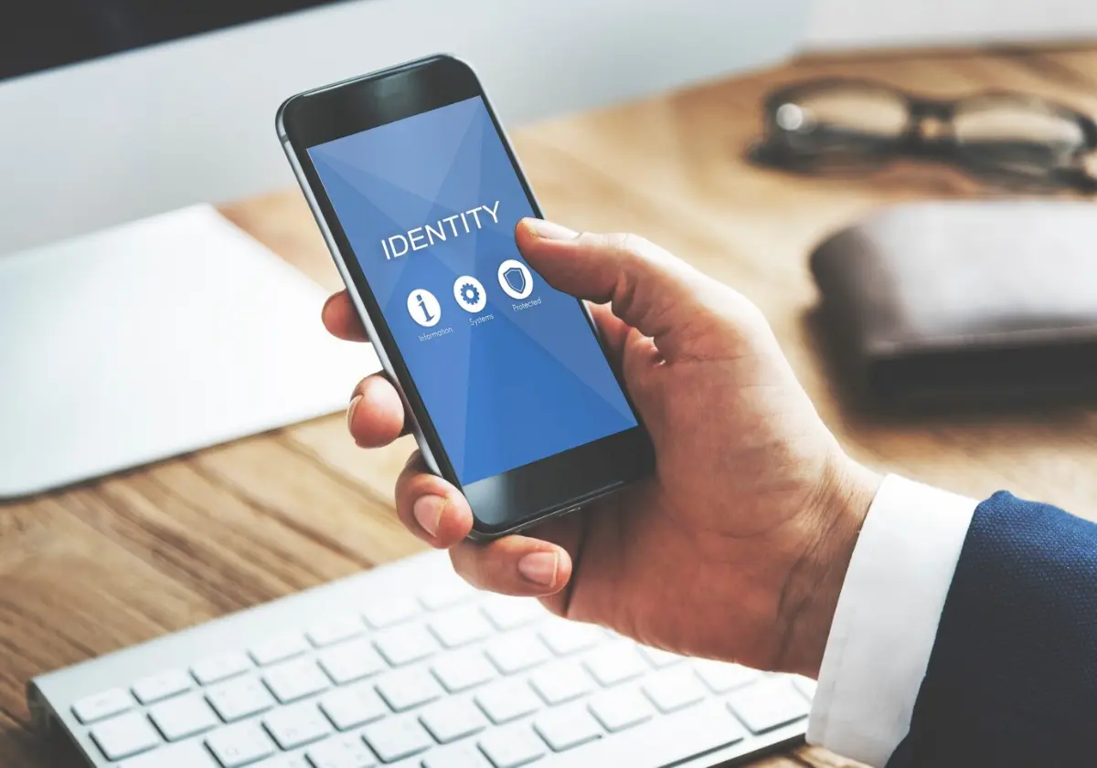 A person holding a smartphone displaying an IDENTITY app, with icons for information, systems, and passwords on a wooden desk.