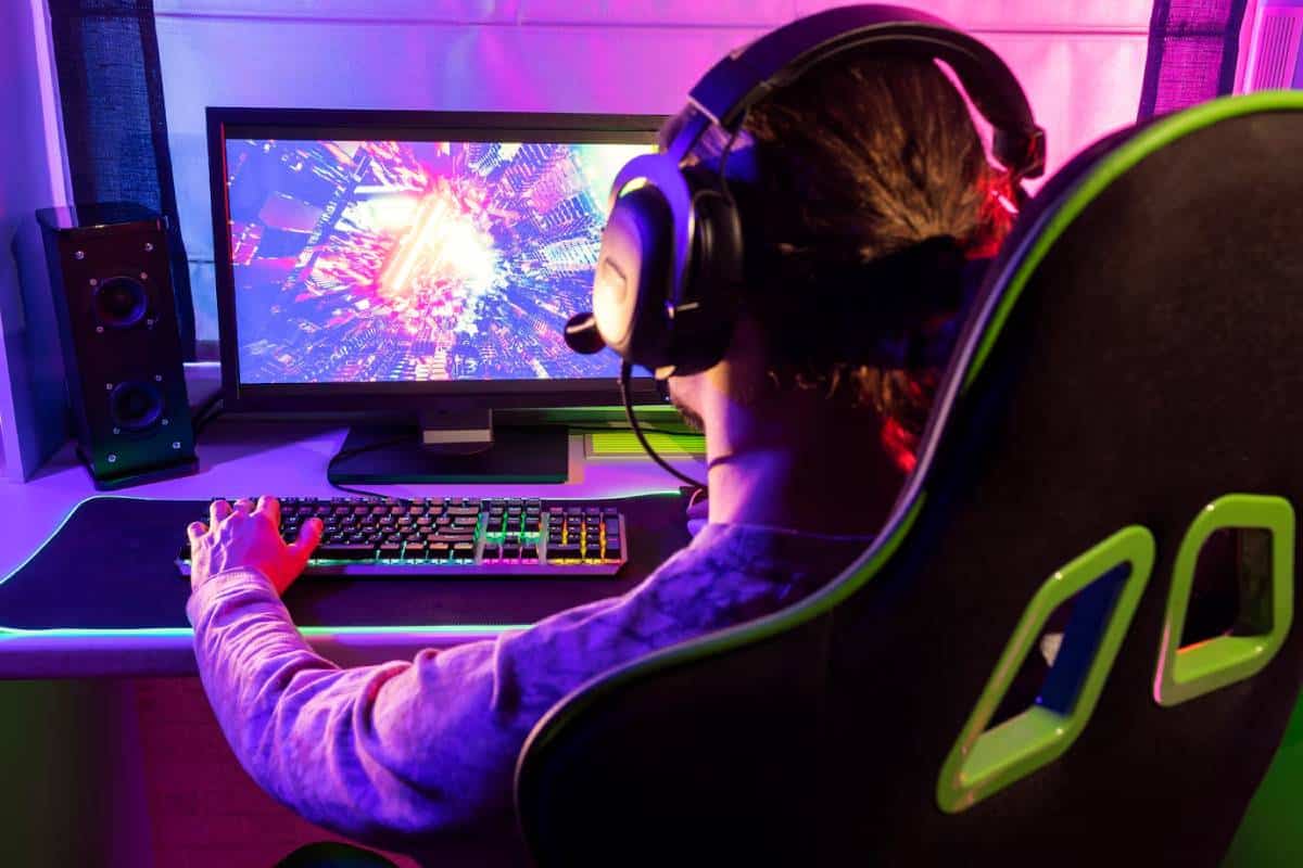 A gamer in a cozy setup wears a headset, engaged with a colorful, dynamic game on the screen, illuminated by ambient lights.