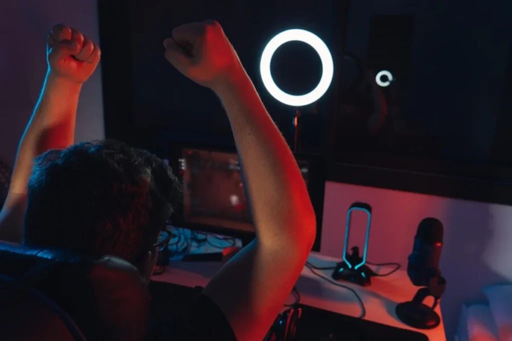 A gamer celebrates a victory, raising clenched fists in excitement, surrounded by glowing lights and a microphone in a dark room.
