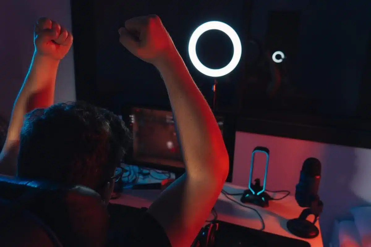 A gamer celebrates a victory, raising clenched fists in excitement, surrounded by glowing lights and a microphone in a dark room.