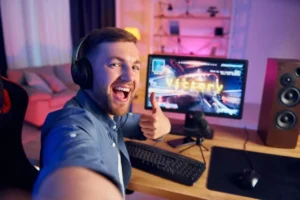 A gamer in a headset gives a thumbs-up, sitting at a desk with a keyboard, mouse, and a monitor displaying "Victory" in bright colors.