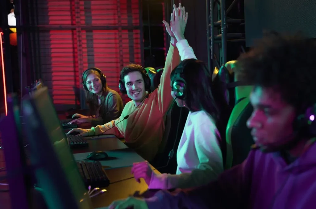 A group of gamers with headsets celebrates a victory, raising their hands in a colorful, illuminated gaming environment.