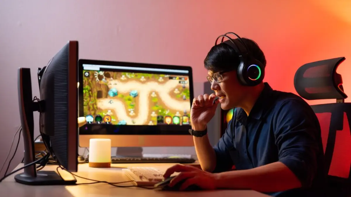 A person gaming at a computer desk, engaged with a vibrant video game displayed on a large monitor, illuminated by soft lighting.