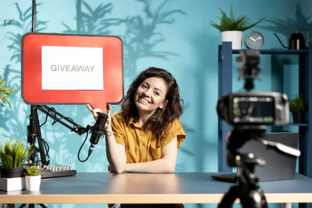 A person in a yellow shirt holds a red sign labeled GIVEAWAY while seated at a desk with a microphone and camera.