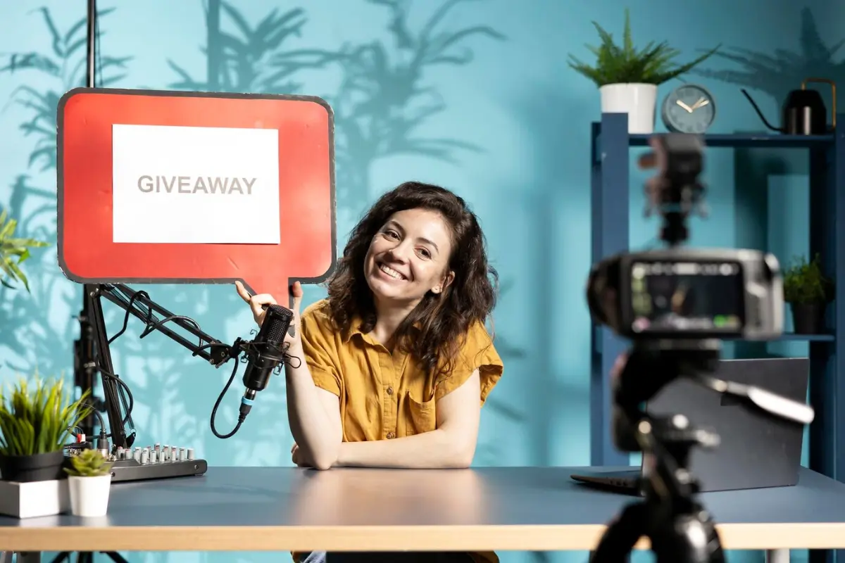 A person in a yellow shirt holds a red sign labeled GIVEAWAY while seated at a desk with a microphone and camera.