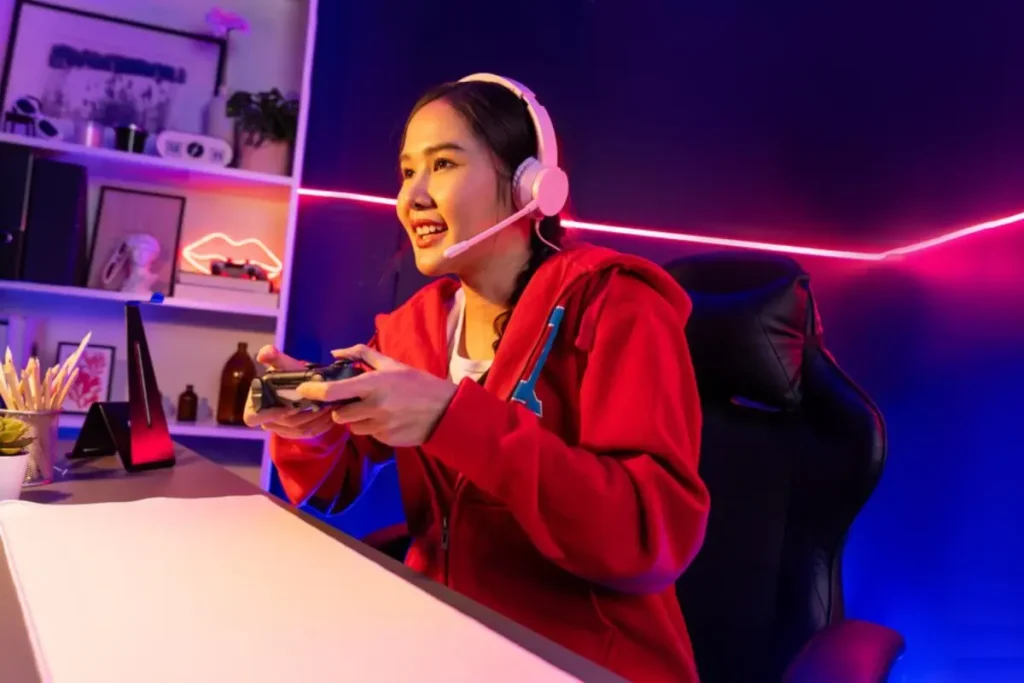 A person wearing a red hoodie and headphones is gaming, holding a controller in a colorful, well-lit room with decorative elements.
