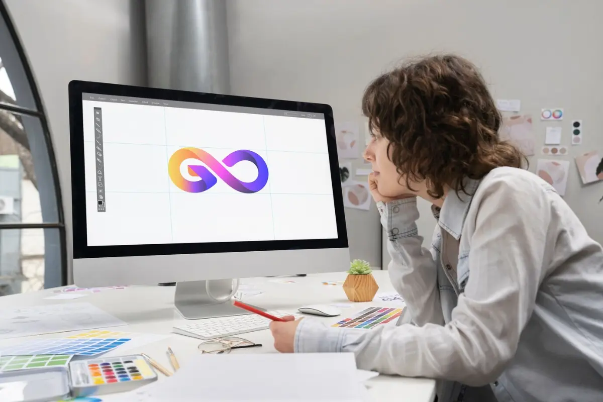 A person sits at a desk, focused on a computer screen displaying a colorful infinity logo, surrounded by design materials.