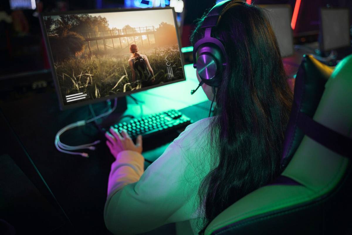 A gamer with long hair wears headphones, focused on a vibrant monitor displaying a character in a lush outdoor scene.