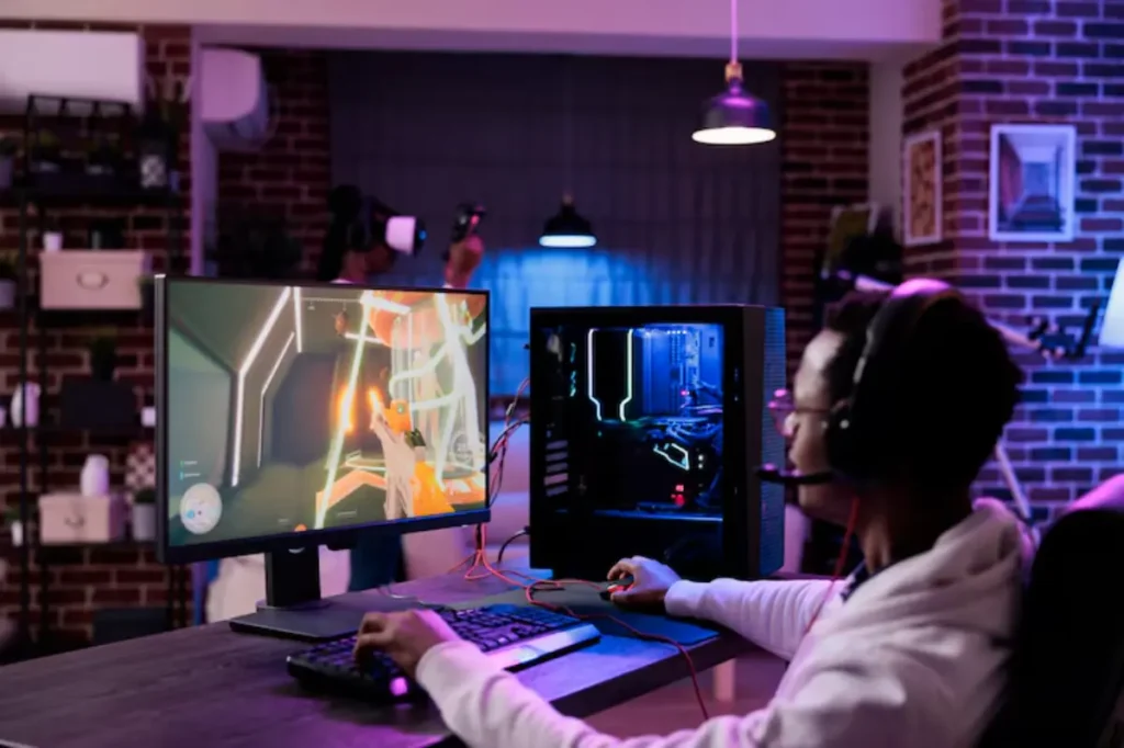 A gamer in a white hoodie plays on a dual monitor setup, focused on a vibrant game, with VR equipment visible in the background.