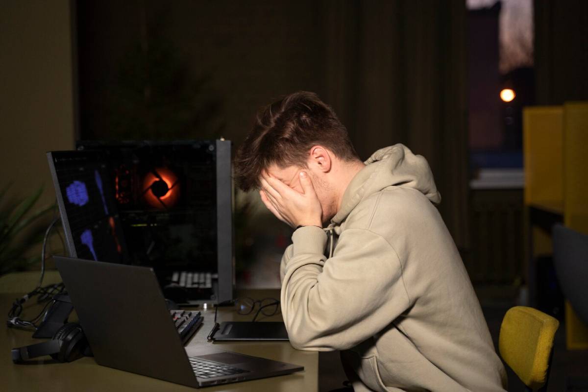 A person in a hoodie sits at a desk, focused on a computer and laptop, with soft lighting and a cozy atmosphere in the background.