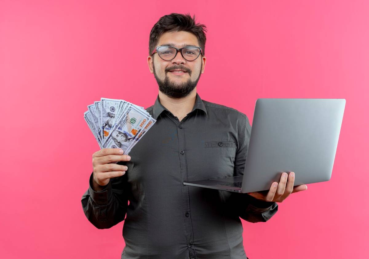 A person in a gray shirt holds a laptop in one hand and a fan of hundred-dollar bills in the other, against a pink background.